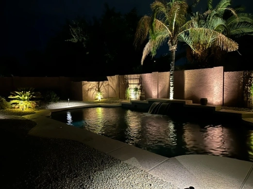 Softly lit palm trees, textured walls, and a pool with a water feature glowing under nighttime landscape lighting.