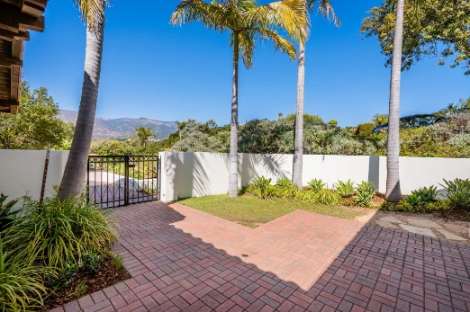 Sunny backyard patio with brick pavers, palm trees, and a white privacy wall; a metal gate to the left and distant hills in view.