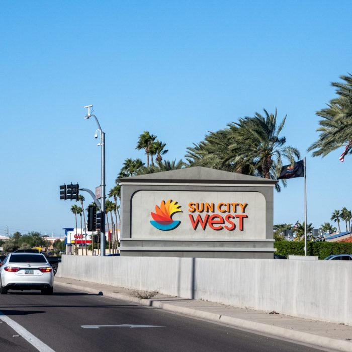 Sun City West entrance sign along Bell Road in Arizona, surrounded by palm trees and landscaped greenery.