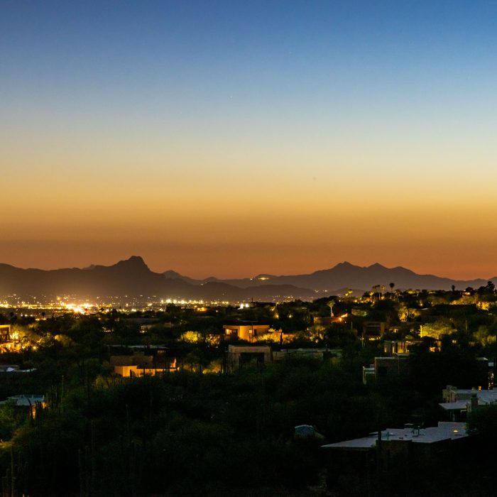 Twilight view of illuminated desert homes and distant city lights beneath a colorful sky and mountain silhouettes.