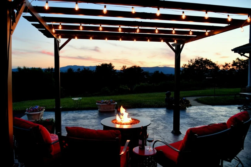 Outdoor Patio Lighting and Fire Pit | Elite Tree Service An outdoor patio at dusk features a lit fire pit table, red-cushioned chairs, and a pergola decorated with string lights overlooking rolling hills.