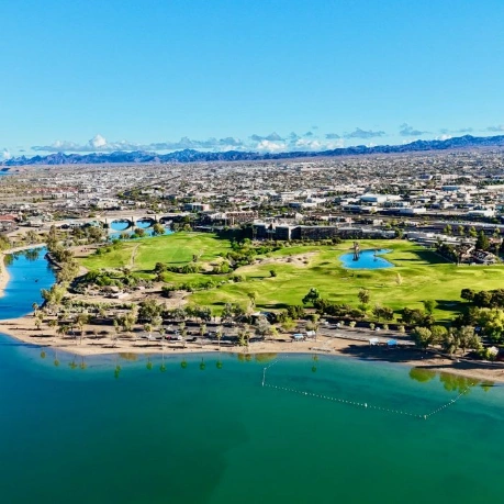 Aerial view of a turquoise lake, sandy beach, and green golf course in Sun Lakes, Arizona, with a city and distant mountains.