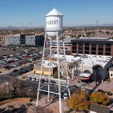 Aerial view of the historic white water tower in downtown Gilbert, Arizona, surrounded by commercial buildings, parking lots, and palm trees.