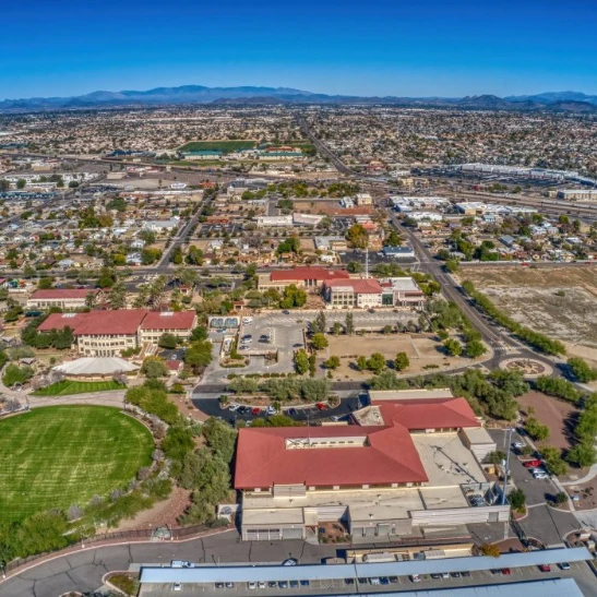 Aerial view of a suburban campus with red-roofed buildings, green fields, and a grid of streets stretching to the city horizon.