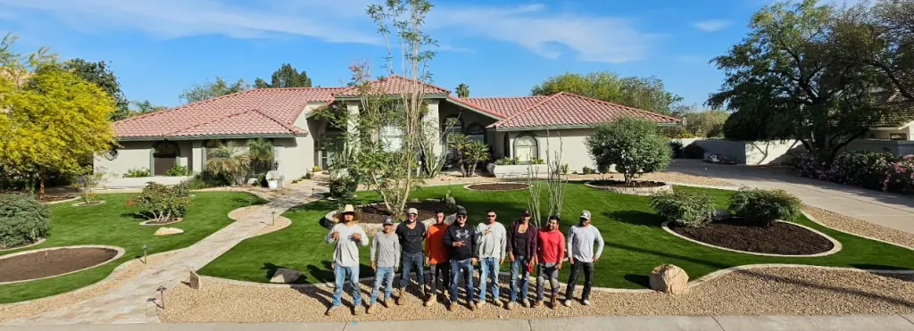 Group of people posing in a sunny front yard in front of a single-story beige house with a red-tiled roof and manicured lawn.