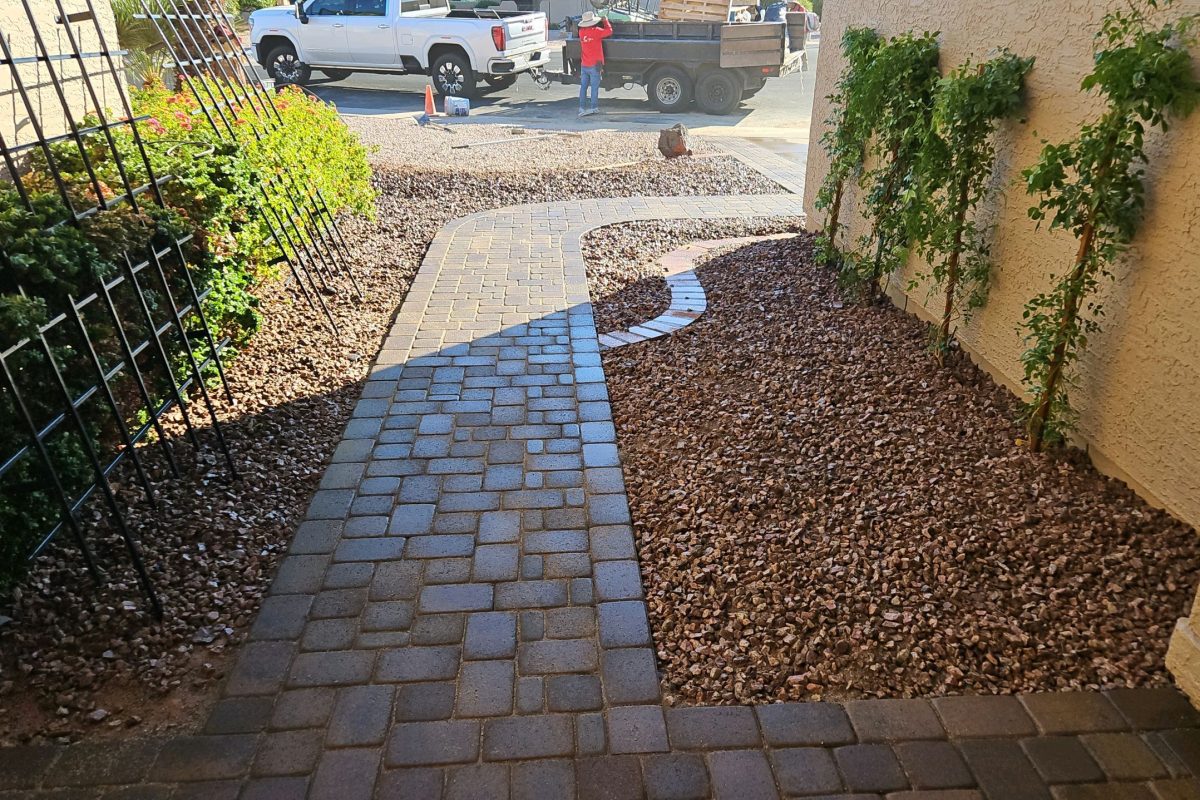 Interlocking stone paver walkway bordered by brown gravel and trellises alongside a residential home.