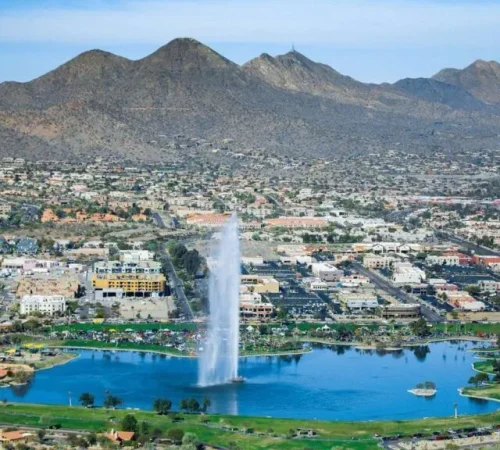 Aerial view of a massive water fountain erupting from a lake in Fountain Hills, Arizona, with a town and mountains in the background.