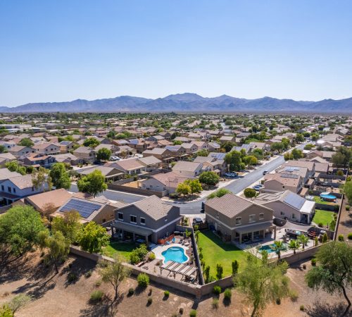 Aerial view of a suburban neighborhood with modern homes, solar panels, and backyard pools in Surprise, Arizona, with mountains in the background.